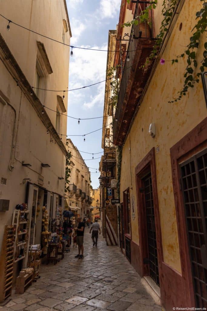 Eine enge Kopfsteinpflasterstraße in einer Altstadt in Apulien, gesäumt von alten Gebäuden. Läden mit Souvenirs und Pflanzen auf Balkonen sind zu sehen - der Charme der Sehenswürdigkeiten in Gallipoli. Zwei Menschen gehen unter Lichterketten und einem teilweise bewölkten Himmel davon.