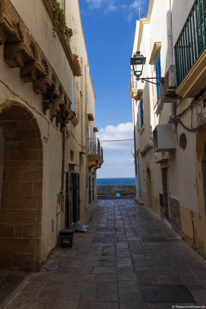 Eine schmale steinerne Gasse zwischen alten beigen Gebäuden führt zu einem Blick auf das blaue Meer unter einem teilweise bewölkten Himmel. Entlang der ruhigen, sonnenbeschienenen Straße sind Balkone, Fenster mit Fensterläden, eine alte Straßenlaterne und eine Mülltonne zu sehen.