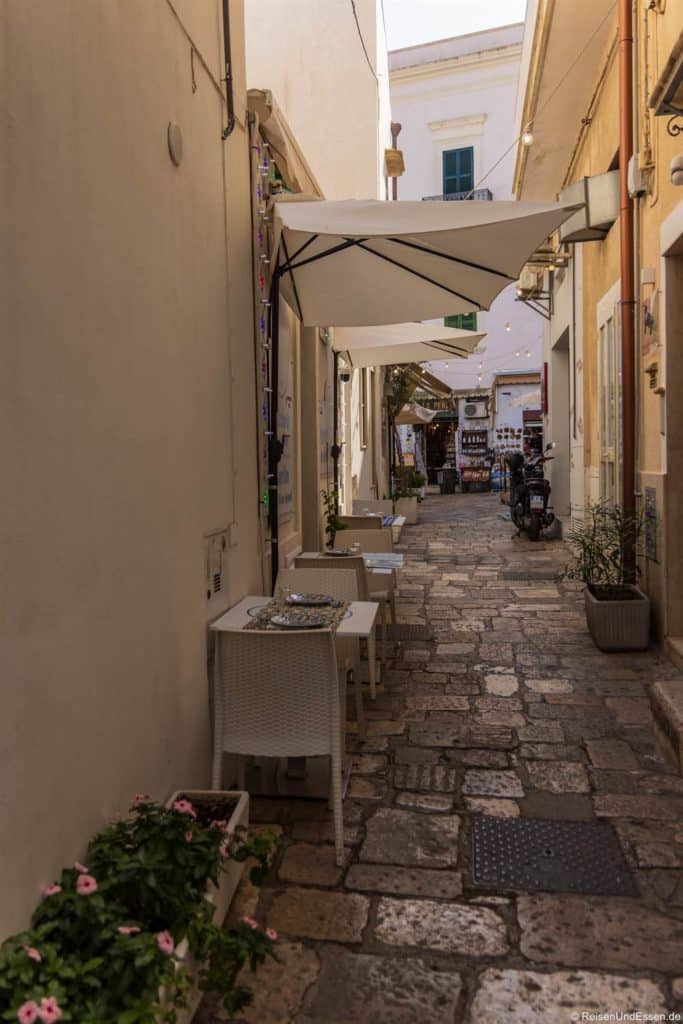 Schmale Kopfsteinpflastergasse mit weißen Wänden und Tischen im Freien unter großen Sonnenschirmen. Topfblumen säumen den Vordergrund. Weiter unten versammeln sich Menschen in der Nähe eines Gallipoli Altstadt Restaurants, während oben Lichterketten im Sonnenlicht hängen.