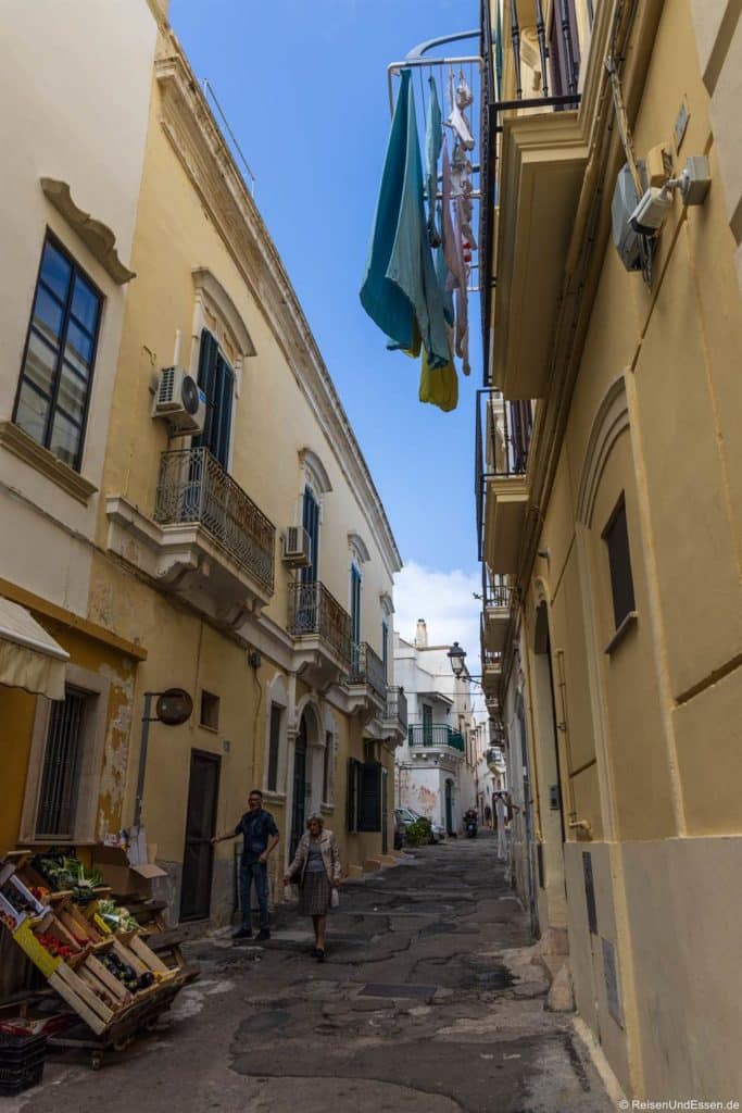 Enge Gasse mit rissigem Pflaster zwischen gelben und weißen Gebäuden in der Altstadt von Gallipoli. Ein Mann und eine Frau gehen an einem hölzernen Einkaufswagen mit Waren vorbei, während über ihnen Wäsche von einem Balkon herabhängt. Der blaue Himmel trägt zum mediterranen Flair bei.