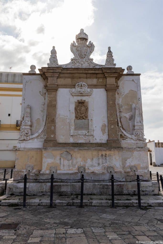 Ein verwittertes, verziertes Steindenkmal in Gallipoli mit barocken Details steht auf einem Platz mit Kopfsteinpflaster. Die Fassade weist ein großes Wappen in der Mitte, dekorative Schnitzereien und sichtbaren abblätternden und verblassten Putz auf. Niedrige schwarze Pfosten säumen den Vordergrund; der Himmel ist bewölkt.