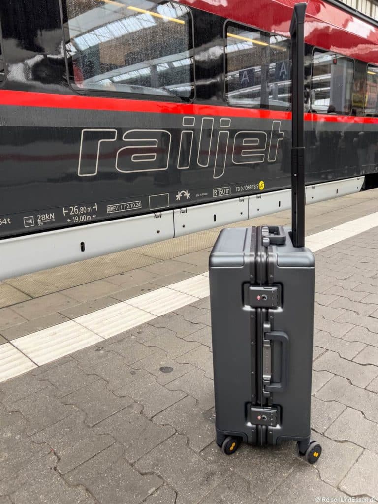Ein grauer Hartschalenkoffer mit verlängertem Griff steht aufrecht auf einem Bahnsteig. Dahinter hält ein moderner rot-schwarzer Railjet-Zug, an dessen Seite das railjet-Logo deutlich zu erkennen ist. Die Oberfläche des Bahnsteigs ist grau und gepflastert.