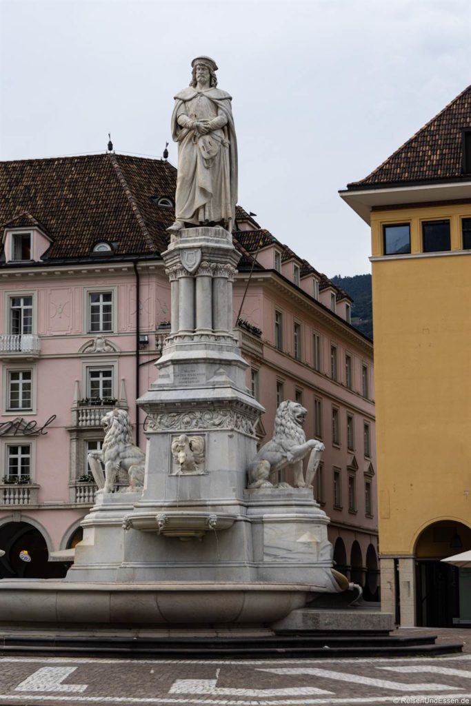 Eine hohe Marmorstatue von Walther von der Vogelweide steht auf einem Sockel, flankiert von zwei sitzenden Löwenskulpturen. Das Denkmal steht auf einem Platz, der von pastellrosa und gelben Gebäuden mit Ziegeldächern und gewölbten Fenstern unter einem wolkenverhangenen Himmel umgeben ist.