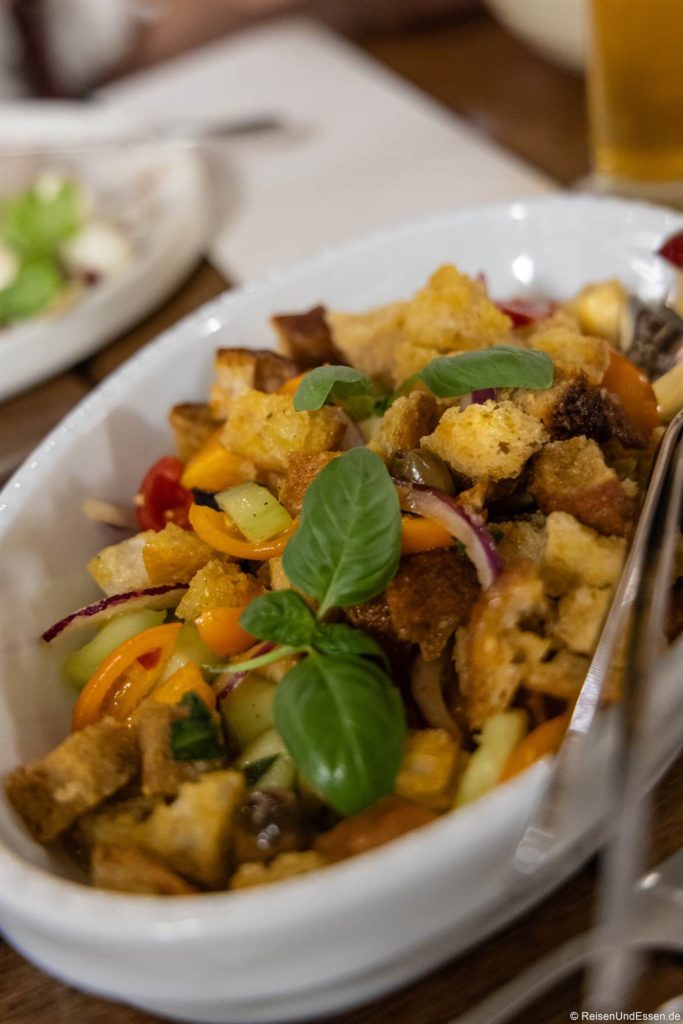 Eine weiße ovale Schale, gefüllt mit buntem Panzanella-Salat mit gerösteten Brotwürfeln, Kirschtomaten, Gurkenscheiben, roten Zwiebeln, gelben Paprikaschoten und frischen Basilikumblättern, mit einer Servierzange an der Seite.
