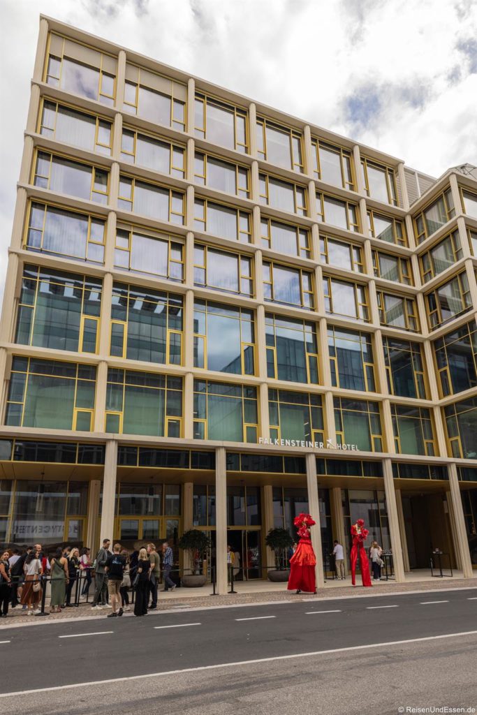 Ein modernes mehrstöckiges Hotel mit großen Glasfenstern und gelben Rahmen. Draußen stehen die Leute Schlange, während in der Nähe des Eingangs zwei Darsteller auf Stelzen in auffälligen roten Kostümen und Kopfbedeckungen stehen. Der Himmel über uns ist teilweise bewölkt.