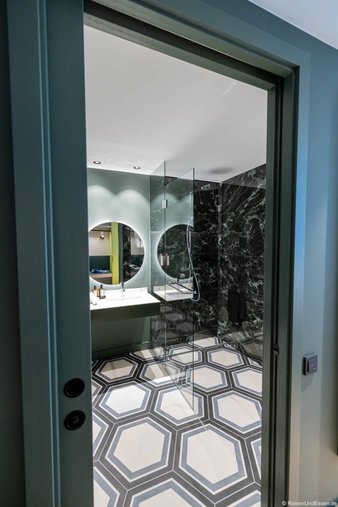 Modernes Badezimmer im Falkensteiner Hotel Bozen, das durch eine offene Tür betrachtet wird, mit einem geometrischen sechseckigen Fliesenboden, grünen Marmorwänden, einer Glasduschabtrennung, einem großen runden Spiegel über einem schwimmenden Waschbecken und einer Deckenbeleuchtung.