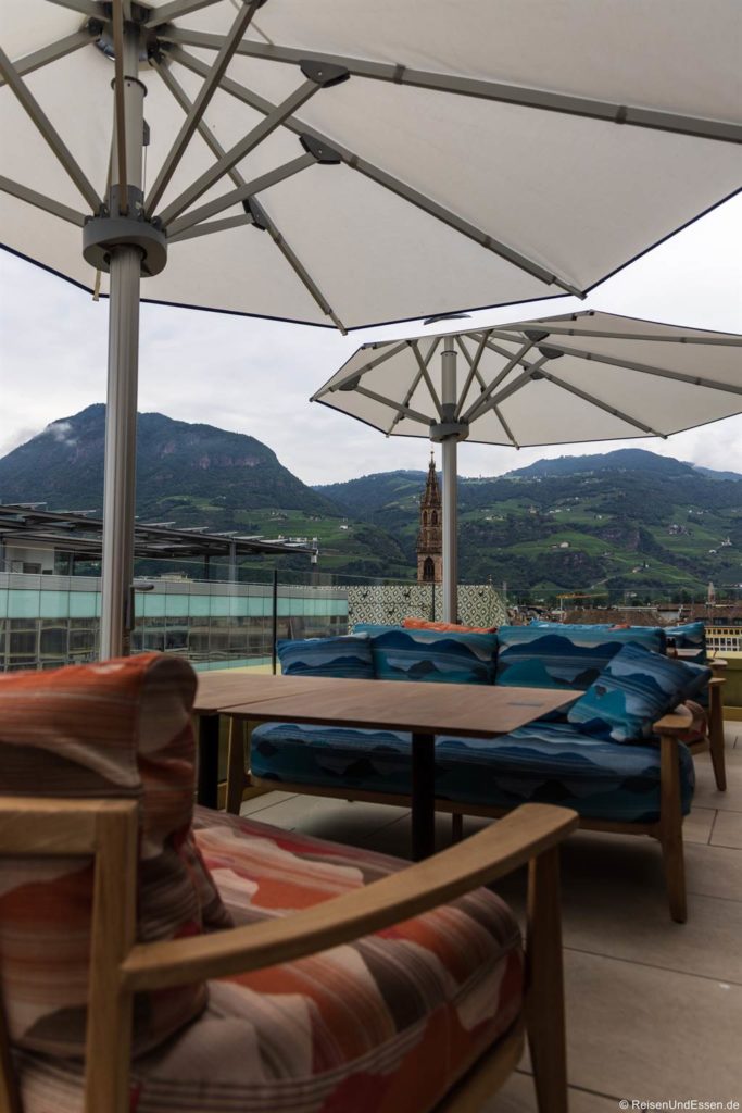Eine Dachterrasse mit gepolsterten Stühlen und einem runden Holztisch unter großen weißen Sonnenschirmen bietet eine malerische Landschaft mit grünen Hügeln, Gebäuden und einem Kirchturm, mit Bergen im Hintergrund unter einem bewölkten Himmel.
