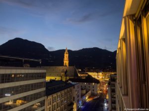 Ein Stadtbild in der Abenddämmerung zeigt Gebäude mit beleuchteten Fenstern, eine Kirche mit einem hohen, beleuchteten Glockenturm in der Mitte und Berge, die sich gegen den tiefblauen Abendhimmel abheben. Die Straßenlaternen leuchten entlang der darunter liegenden Straßen. Die Aussicht vom Falkensteiner Hotel Bozen