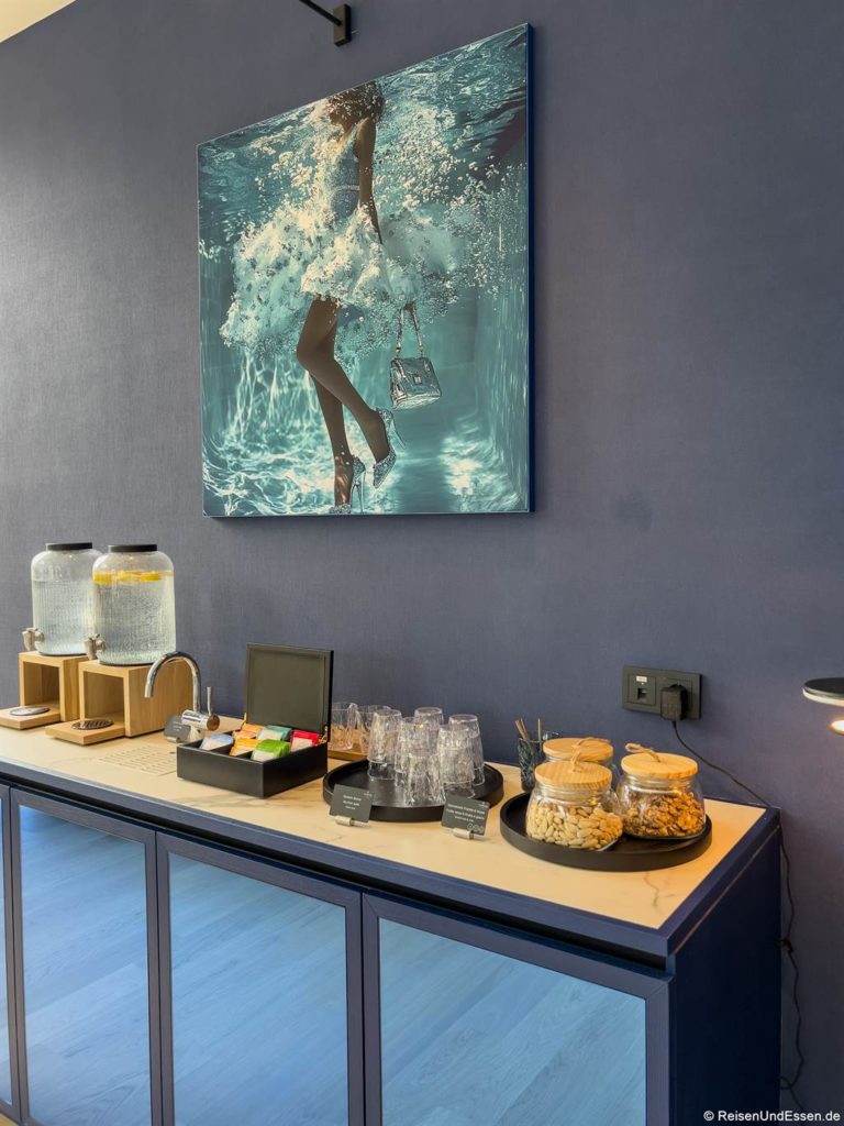 Auf einem blauen Schrank stehen Gläser mit Snacks, gestapelte Gläser, Teebeutel und zwei Spender für Wasser und Limonade. Darüber befindet sich ein Fotokunstwerk, das eine Frau in einem weißen Kleid und hochhackigen Schuhen unter Wasser zeigt, die eine Handtasche hält. Die Wand und der Schrank sind beide blau.