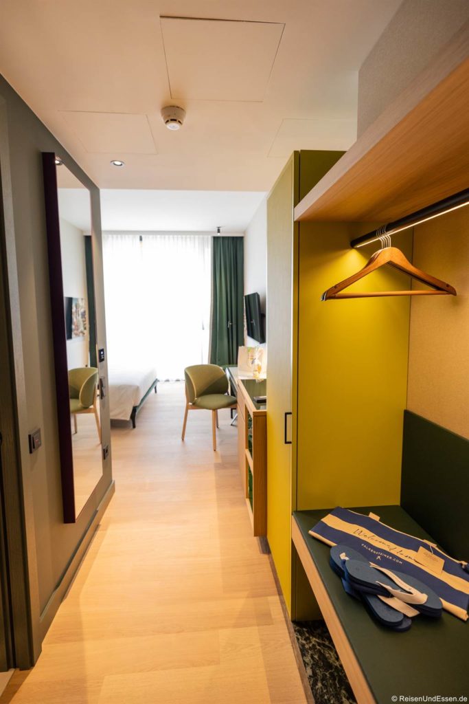 Helles, modernes Hotelzimmer im Falkensteiner Hotel Bozen mit hellem Holzboden, grüner Akzentwand, offenem Kleiderschrank mit Kleiderbügeln, Schreibtisch, grünem Stuhl und großem Fenster mit weißen Vorhängen, die das Tageslicht hereinlassen. Eine blaue Reisetasche liegt auf der Bank neben dem Eingang.