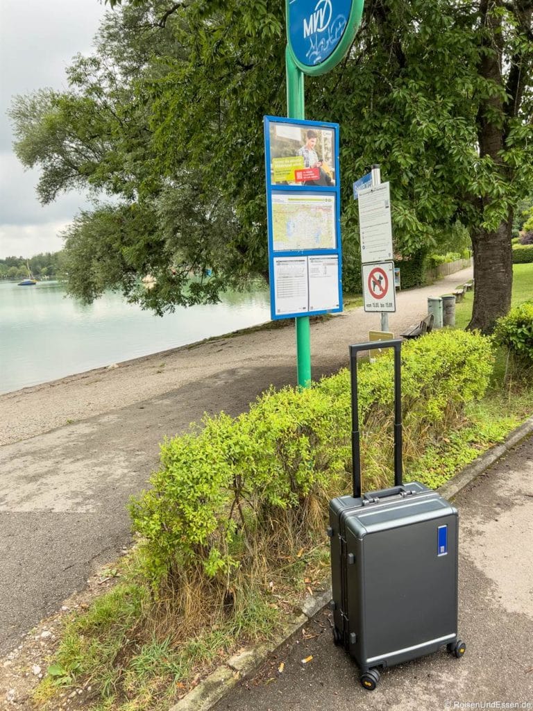 Ein geschlossener schwarzer Koffer mit Rädern und einem verlängerten Griff steht auf einem gepflasterten Weg neben niedrigen grünen Sträuchern. Dahinter befinden sich ein See, Bäume und mehrere Schilder an einem Pfahl, darunter eine Karte und ein Verbotsschild für Hunde. Der Himmel ist bedeckt.