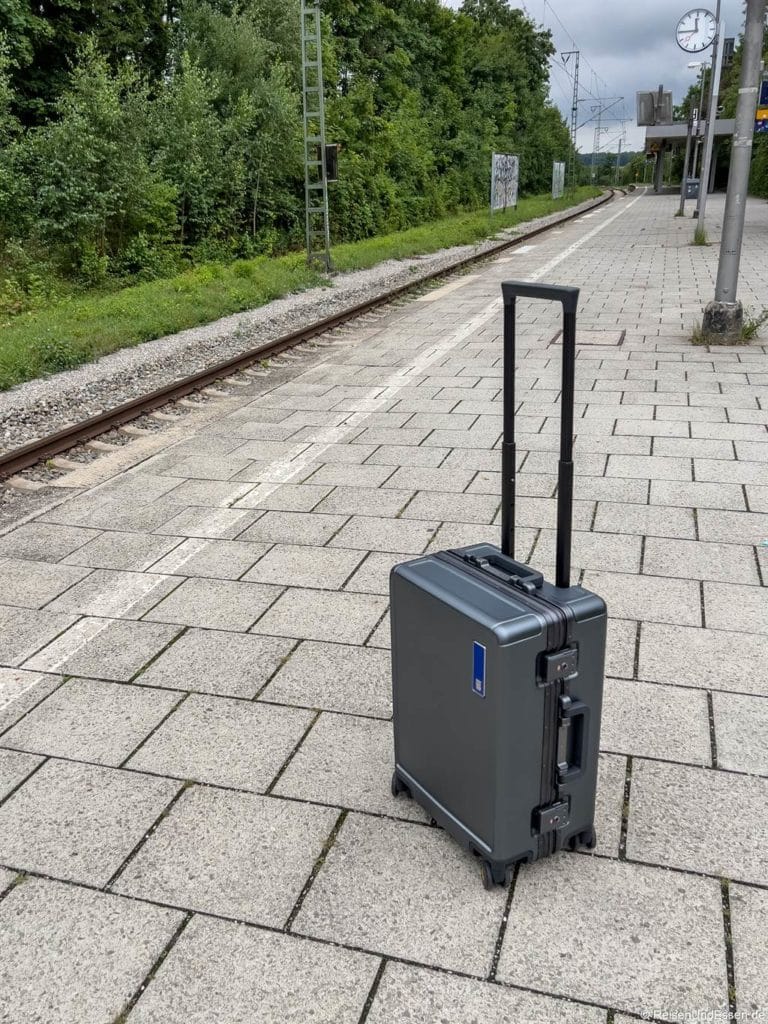 Ein einzelner grauer Rollkoffer mit einem verlängerten Griff steht auf einem gepflasterten, leeren Bahnsteig neben den Gleisen. Bäume säumen den Hintergrund, und eine Bahnhofsuhr zeigt bei bewölktem Himmel etwa 12:10 Uhr an.