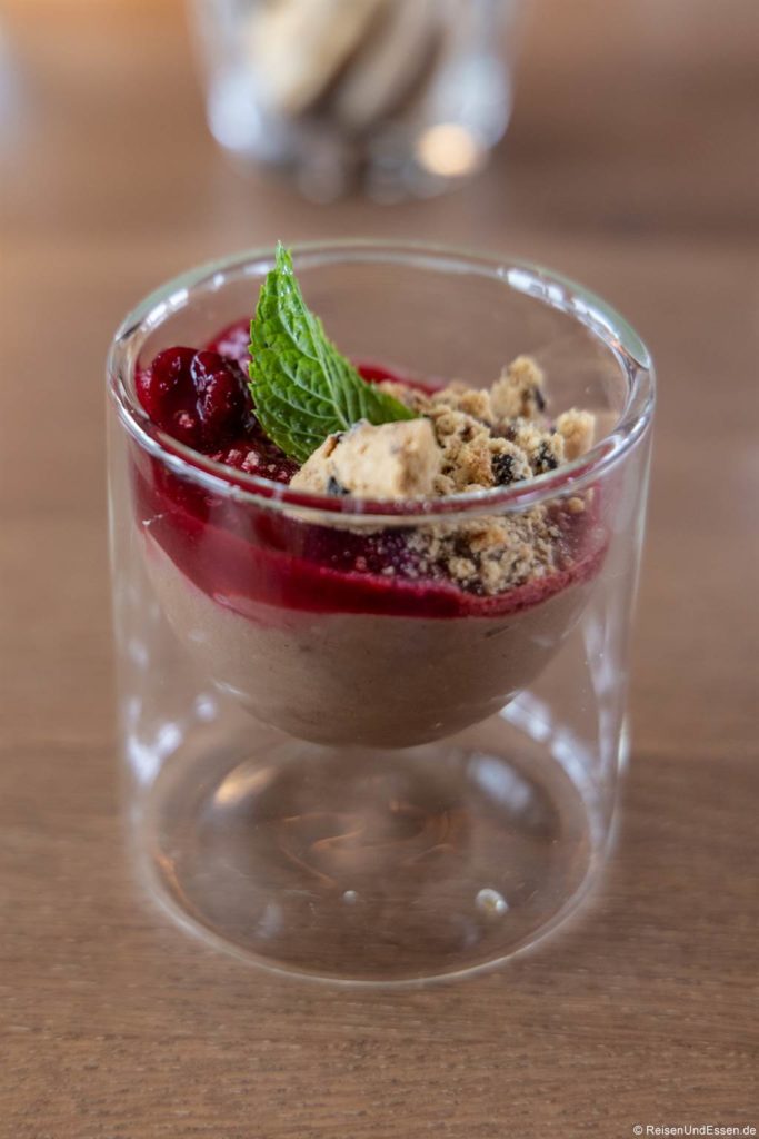 Auf einer Holzplatte steht ein Glas mit Mousse au Chocolat, das mit roter Beerensauce, zerbröselten Keksen und einem frischen Minzblatt garniert ist. Die Mousse hat eine weiche Textur, und das Dessert wird in einem doppelwandigen Glas serviert.