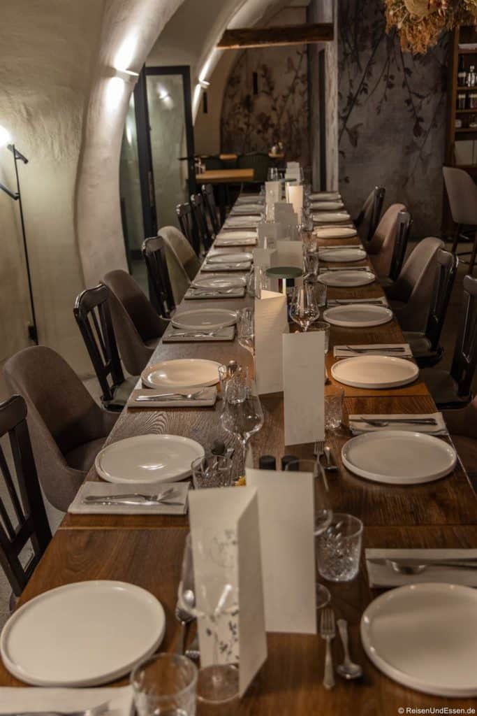 Ein langer, eleganter Esstisch im Restaurant Bogen in Bozen, gedeckt für eine Mahlzeit in einem schwach beleuchteten Raum mit Gewölbedecken. Auf jedem Platz stehen ein weißer Teller, ein Glas, Besteck und eine gefaltete Speisekarte. Dunkle Holzstühle säumen den Tisch, und die Atmosphäre ist gemütlich und einladend.