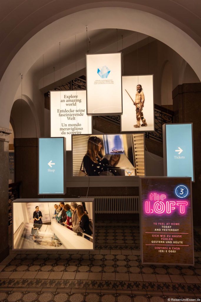 Eine Museumslobby mit beleuchteten Hängeschildern in Englisch und Deutsch, darunter "Explore an amazing world", Bilder eines prähistorischen Menschen, Besucher, die mit Exponaten interagieren, Shop- und Ticketschilder und ein Neonschild für "the Loft". Im Hintergrund befinden sich verzierte Treppen und Bögen.