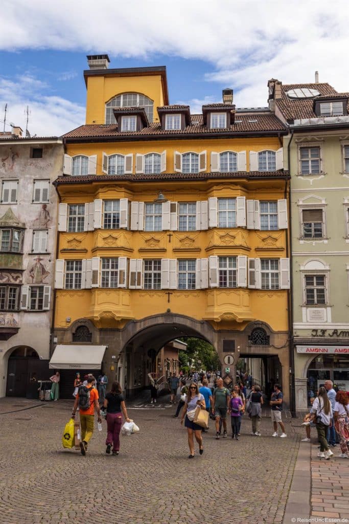 Ein belebter Platz mit Kopfsteinpflaster und Menschen, die vor einem hohen, gelben Gebäude mit weißen Fensterläden, gewölbten Türen und einem Durchgang darunter spazieren gehen. Die angrenzenden Gebäude sind blassgrün und beige, und der Himmel über ihnen ist teilweise bewölkt.