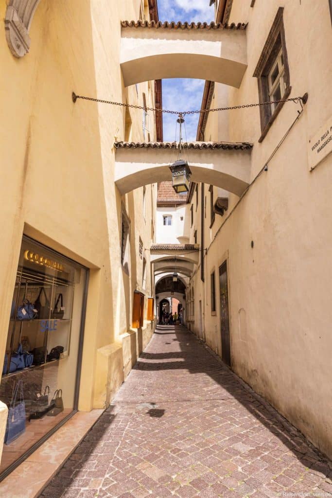 Eine schmale Gasse mit Kopfsteinpflaster zwischen gelben und beigen Gebäuden mit gewölbten Übergängen, Fenstern und einer hängenden Laterne. Auf der linken Seite zeigt ein Schaufenster Taschen und ein Verkaufsschild. In der schattigen Ferne laufen Menschen unter weiteren Bögen.