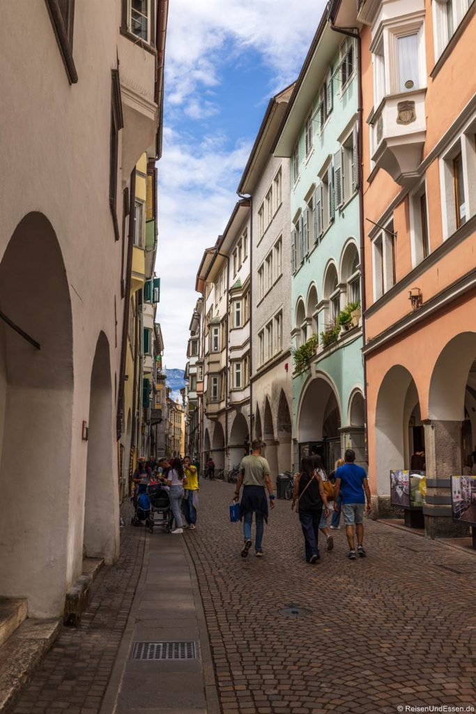 Eine Straße mit Kopfsteinpflaster, gesäumt von bunten, hohen Gebäuden mit Bögen und Balkonen. Die Menschen flanieren und sitzen vor den Cafés unter einem teilweise bewölkten Himmel. Die Szene ist lebendig, mit einer Mischung aus Fußgängern und Sitzgelegenheiten im Freien, die eine charmante europäische Stadtatmosphäre hervorrufen. Die Laubengasse gehört zu den Sehenswürdigkeiten in Bozen.
