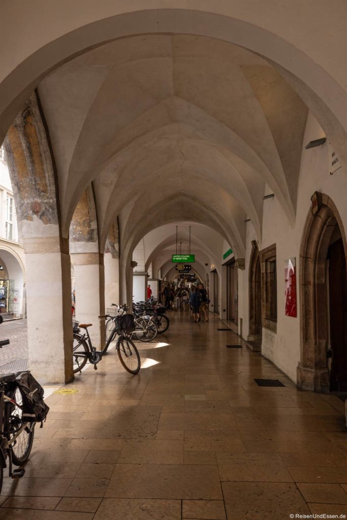 Eine gewölbte Steinpassage in der Laubengasse, die zu den Sehenswürdigkeiten in Bozen zählt, mit gotischen Bögen, Fahrrädern, die an der linken Wand abgestellt sind, beigem Kachelboden und Menschen, die in der Ferne spazieren gehen. Das Sonnenlicht fällt durch die Bögen, und ein grünes Schild hängt in der Mitte des Bildes über dem Kopf.