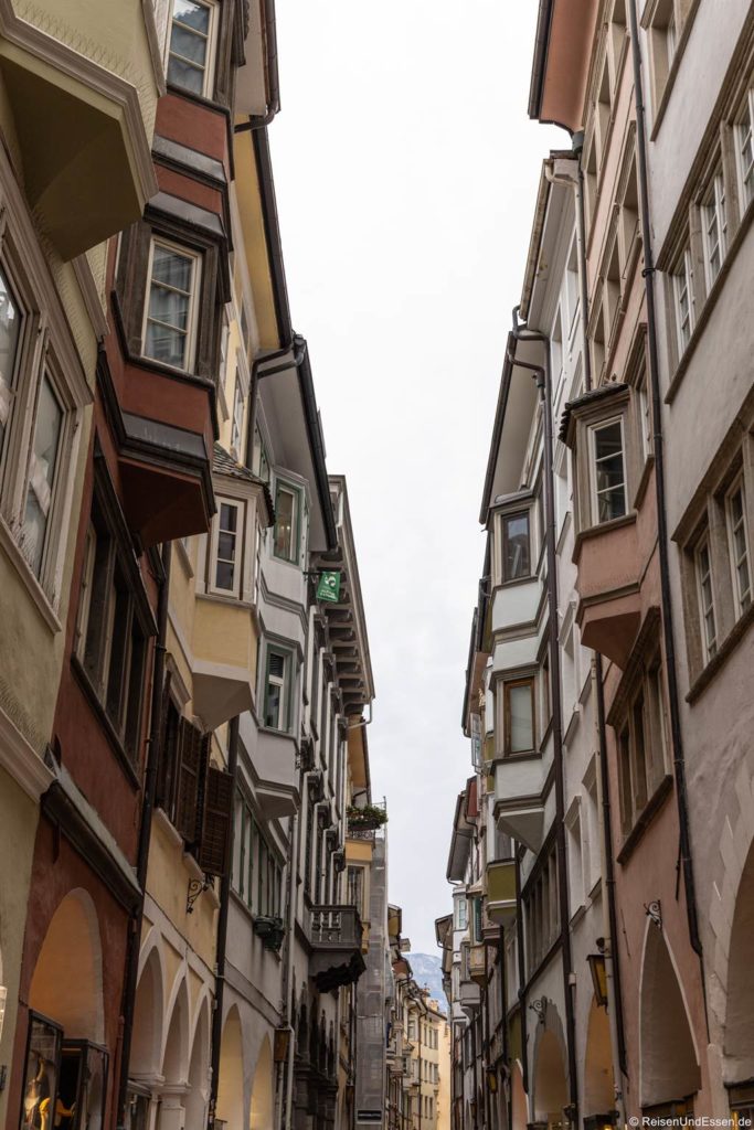 Schmale Straße mit hohen, farbenfrohen Gebäuden mit überhängenden Erkerfenstern und Bögen. Die Gebäude stehen dicht beieinander, so dass unter dem wolkenverhangenen Himmel ein canyonartiger Effekt entsteht. Die Perspektive lenkt den Blick auf die weit entfernte Stadt dahinter. Die Laubengasse gehört zu den Sehenswürdigkeiten in Bozen