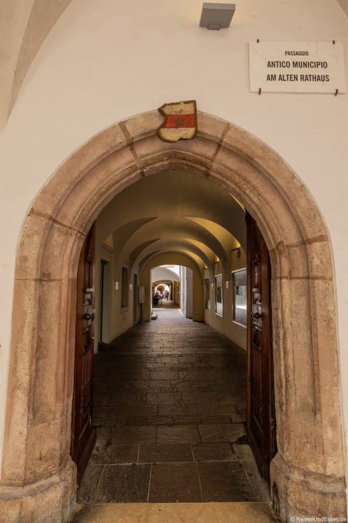 Ein steinerner Torbogen mit Holztüren führt in einen gewölbten Korridor, der mit Bögen und Lichtern versehen ist. Über dem Bogen befindet sich ein Wappen und ein Schild mit der Aufschrift "Passaggio Antico Municipio am Alten Rathaus". Der Gang führt zu einem hellen Ausgang am anderen Ende.
