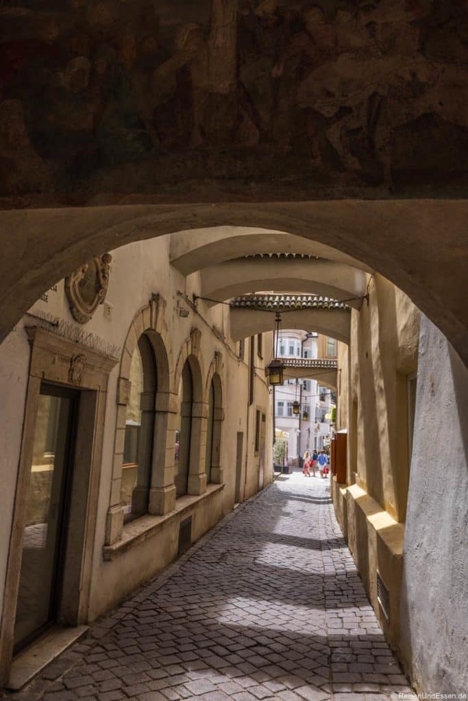 Eine enge Gasse mit Kopfsteinpflaster, gewölbten Steinbrücken und gelben Wänden führt zu einer sonnenbeschienenen Straße, auf der sich in der Ferne Menschen versammeln. Weiches Licht und Schatten schaffen eine warme, einladende Atmosphäre in dieser historischen europäischen Stadtszene.