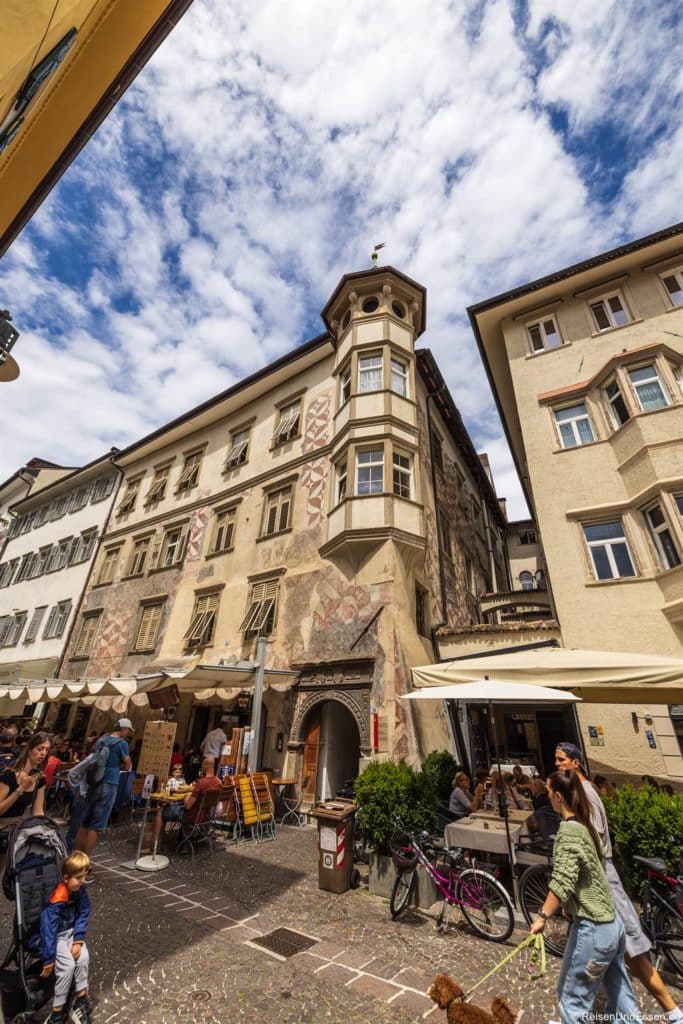 Eine belebte europäische Straßenszene zeigt Menschen an Cafétischen vor einem verzierten, historischen Gebäude mit Wandmalereien und einem Türmchen unter einem teilweise bewölkten Himmel. In der Nähe sind Fahrräder geparkt, und Markisen spenden Schatten auf der kopfsteingepflasterten Straße.