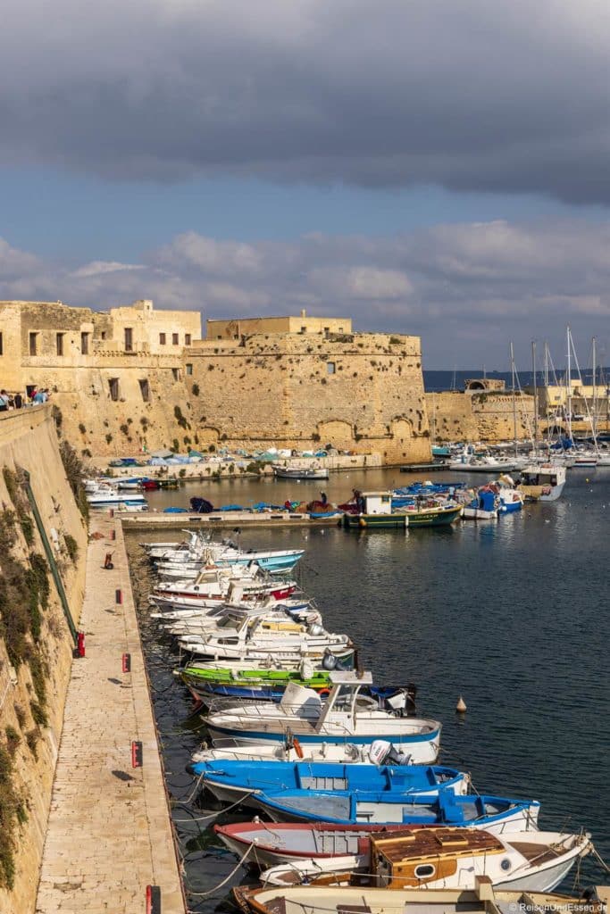 Steinerne Festungsmauern in Gallipoli begrenzen einen Hafen, in dem kleine, teils bunte Boote nebeneinander vertäut sind. Ein steinerner Steg führt am Wasser entlang. Der Himmel ist teilweise bewölkt, und in der Ferne sind Segelboote in der Nähe der Festung im Hintergrund zu sehen.