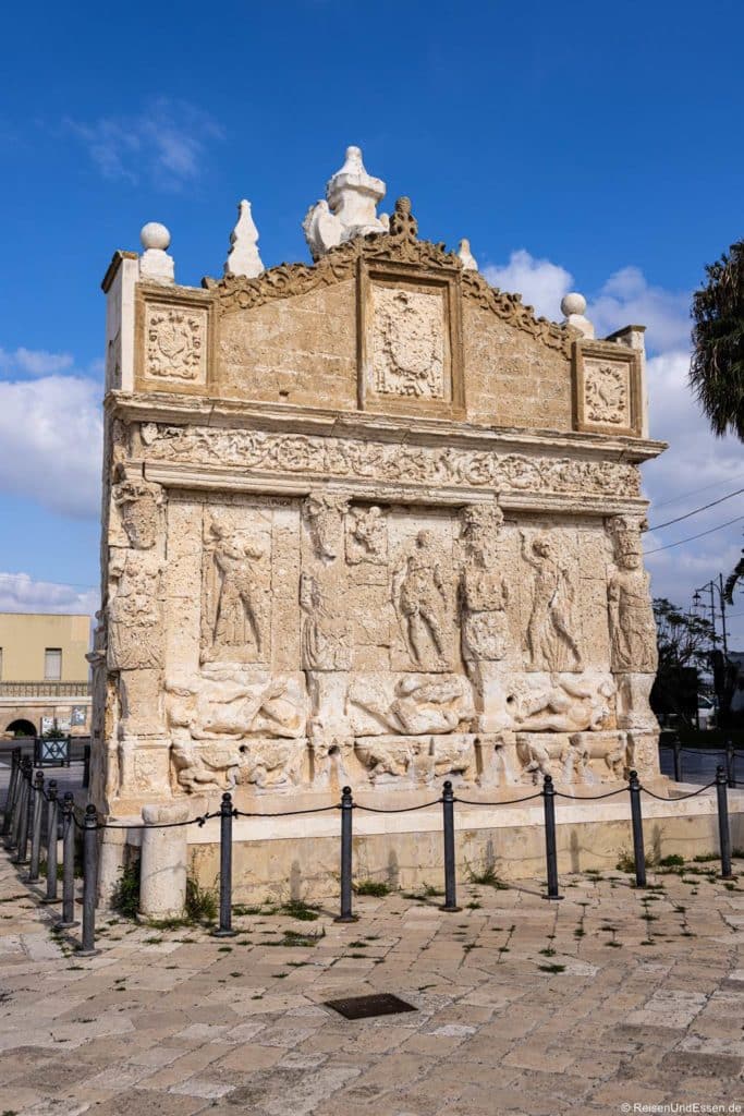 Ein großes Steinmonument mit detaillierten Schnitzereien von menschlichen Figuren, Tieren und dekorativen Mustern steht im Freien auf einer gepflasterten Fläche. Die Struktur weist kunstvolle Reliefs und skulpturale Elemente unter einem blauen Himmel mit vereinzelten Wolken auf. Metallpfosten mit Ketten umgeben das Denkmal.