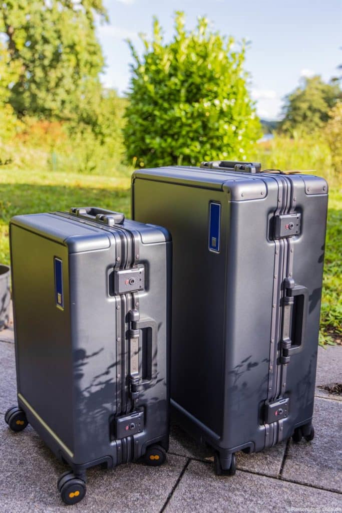 Zwei dunkelgraue Hartschalenkoffer beim Koffertest. Level 8 Hegent Carry-On und Check-In, ein kleiner und ein großer, stehen aufrecht auf einer gepflasterten Fläche im Freien. Beide haben vier Räder und eingebaute Schlösser. Im hellen, sonnenbeschienenen Hintergrund sind üppige grüne Büsche und Bäume zu sehen.