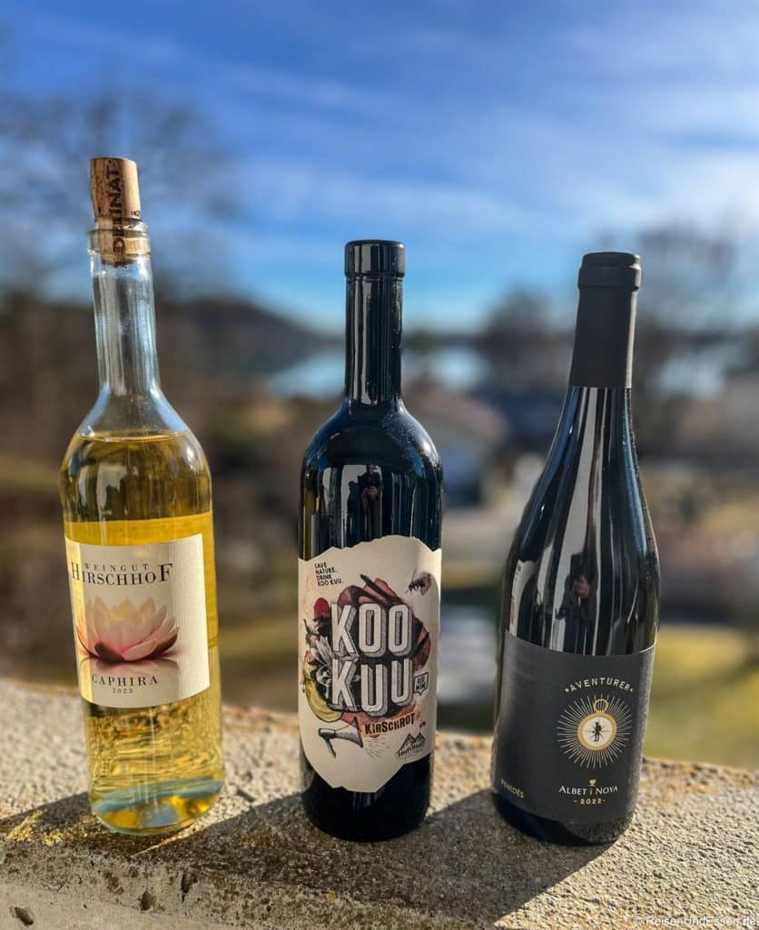 Drei Weinflaschen von Delimat Piwi stehen auf einem Steinsockel im Freien. Von links nach rechts: ein heller, goldener Wein mit weißem Etikett, ein dunkler Wein mit der Aufschrift "Kooku" und ein dunkler Wein mit einem schwarzen Etikett. Im Hintergrund sind eine verschwommene Landschaft und ein blauer Himmel zu sehen.