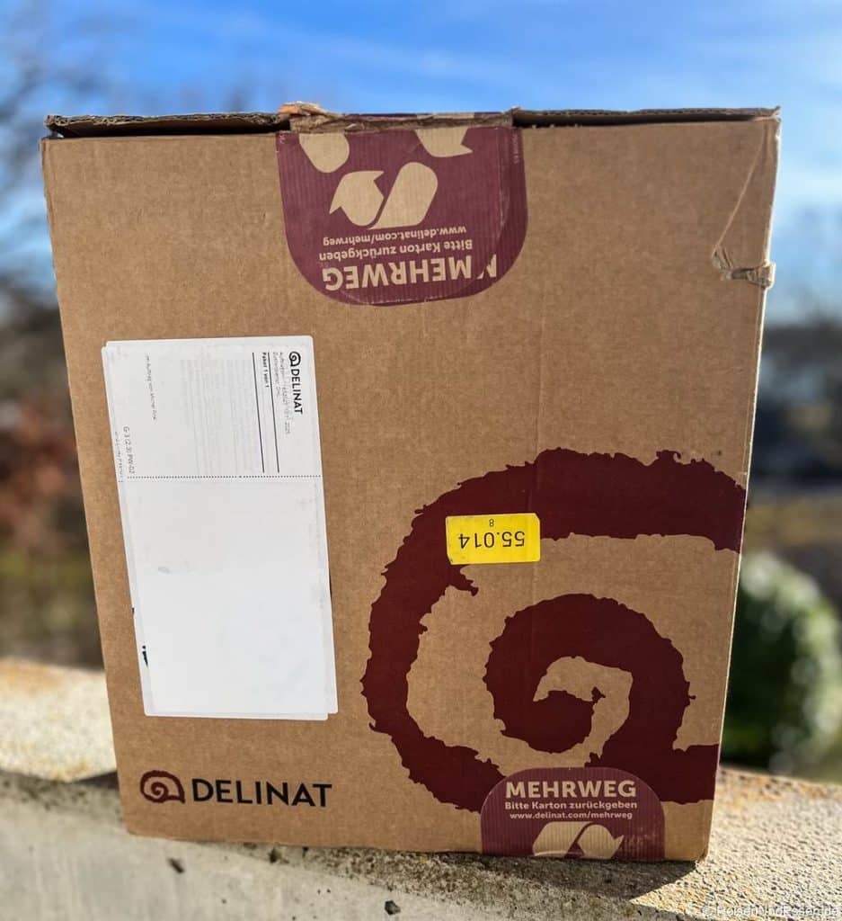 Eine braune Pappschachtel von Delinat steht auf einer Steinfläche im Freien. Auf dem Karton sind ein weinrotes Schneckenlogo und das Wort Mehrweg zu sehen. Im Hintergrund sind Bäume und ein blauer Himmel verschwommen zu sehen.