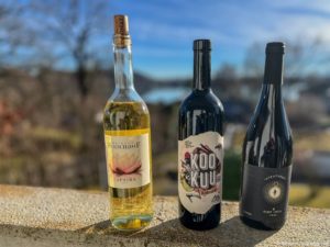 Drei Weinflaschen von Delinat Piwi stehen auf einem Steinsockel im Freien. Die linke Flasche ist klar mit hellgelbem Wein und einem Lotus-Etikett, die mittlere ist dunkel mit einem modernen "Kooku"-Etikett, und die rechte ist dunkel mit einem minimalistischen Etikett, alles vor einem verschwommenen natürlichen Hintergrund.