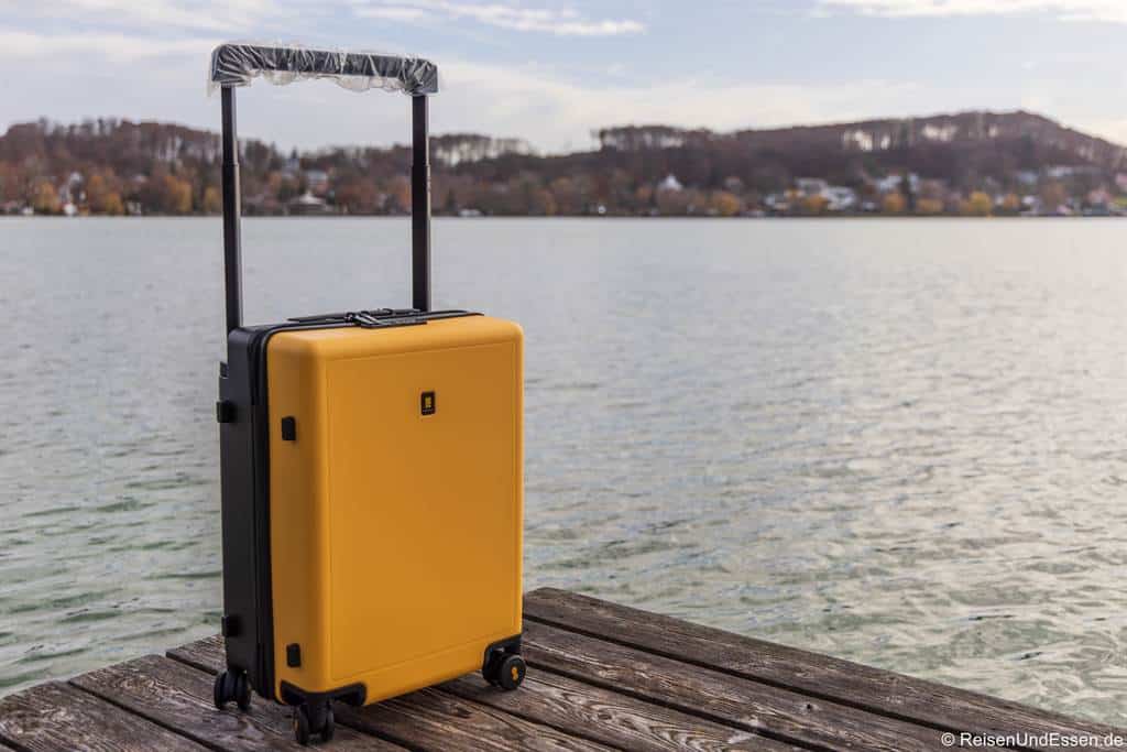 Handgepäck-Koffer für Kurzreisen: Ein gelber Hartschalenkoffer mit schwarzem Griff und Rädern steht auf einem Holzsteg an einem ruhigen See. Im Hintergrund sind entfernte Hügel, Herbstbäume und verstreute Häuser unter einem teilweise bewölkten Himmel zu sehen.