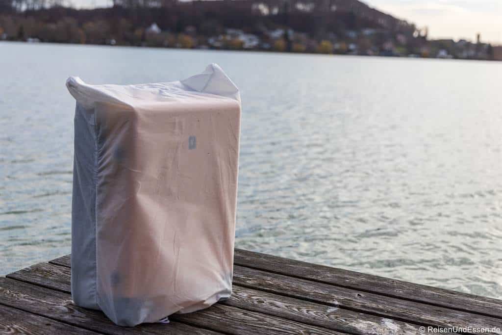 Ein in eine durchsichtige Flieshülle eingewickelter Koffer liegt auf einem Holzsteg an einem ruhigen See. Im Hintergrund sind verschwommene Häuser und ein bewaldeter Hügel unter einem wolkenverhangenen Himmel zu sehen. Die Umgebung wirkt friedlich und leicht bewölkt.