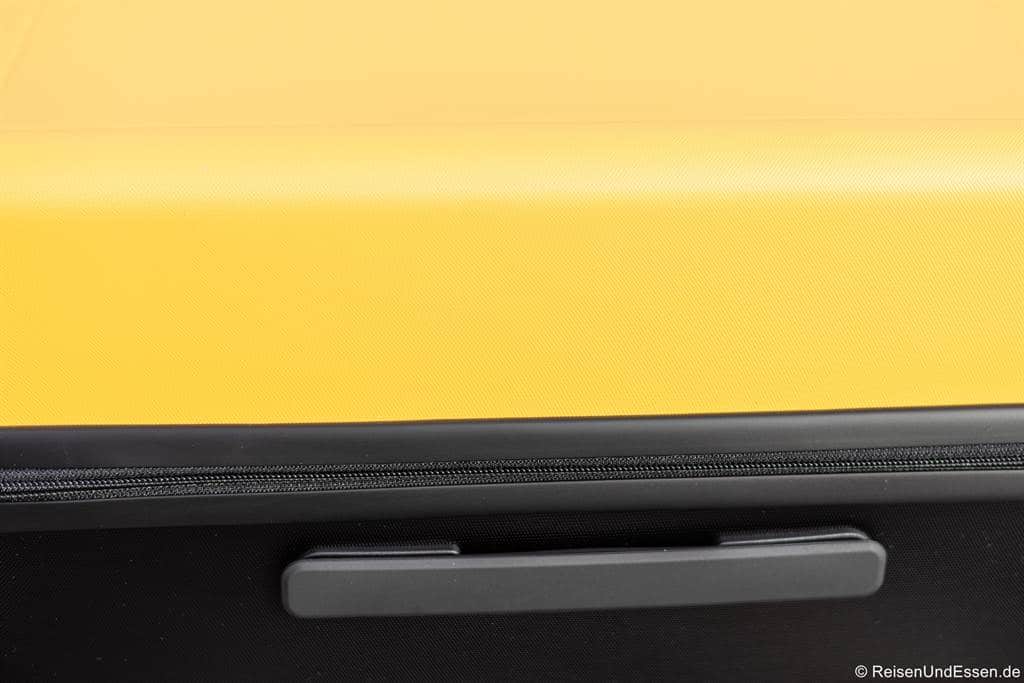 Nahaufnahme eines gelben Hartschalenkoffers, die die strukturierte Oberfläche, einen schwarzen Reißverschluss und einen grauen Kunststoffgriff auf einem schwarzen Teil zeigt. Das Foto ist minimalistisch mit scharfem Farbkontrast, und in der rechten unteren Ecke ist ein kleines Wasserzeichen zu sehen.