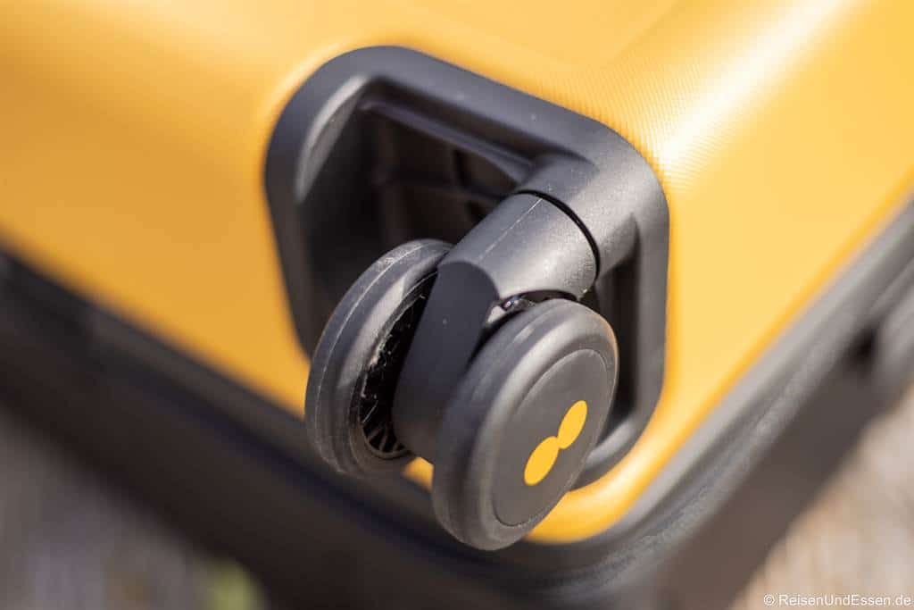 Nahaufnahme eines schwarzen Doppelrads auf einem gelben Hartschalenkoffer beim Level8 Voyageur. Die Oberfläche des Koffers ist strukturiert, und die Räder sind mit einer stabilen schwarzen Kunststoffhalterung befestigt. Das Sonnenlicht hebt die leuchtend gelbe Farbe und das glatte Design des Koffers und der Räder hervor.
