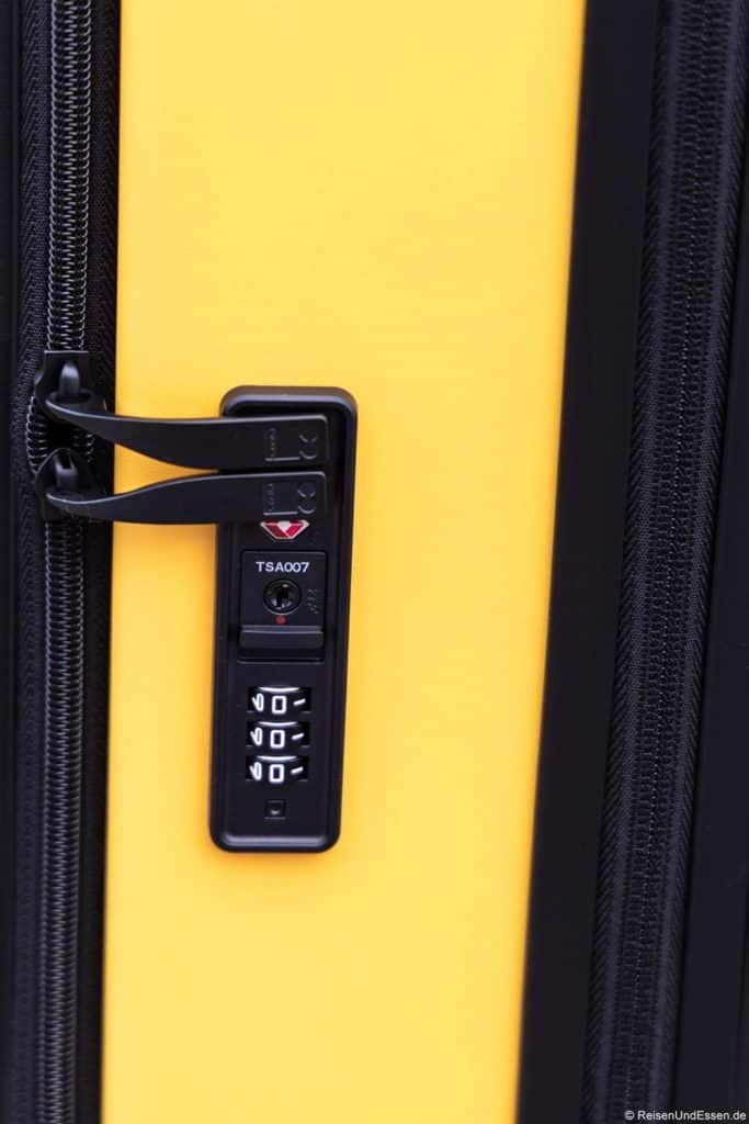 Nahaufnahme eines gelben Koffers mit einem schwarzen Reißverschluss und einem eingebauten TSA-Kombinationsschloss. Das Schloss verfügt über drei Einstellräder, die auf 0-9 eingestellt sind, ein kleines Schlüsselloch darüber und in den Schließmechanismus integrierte Reißverschlusszüge.