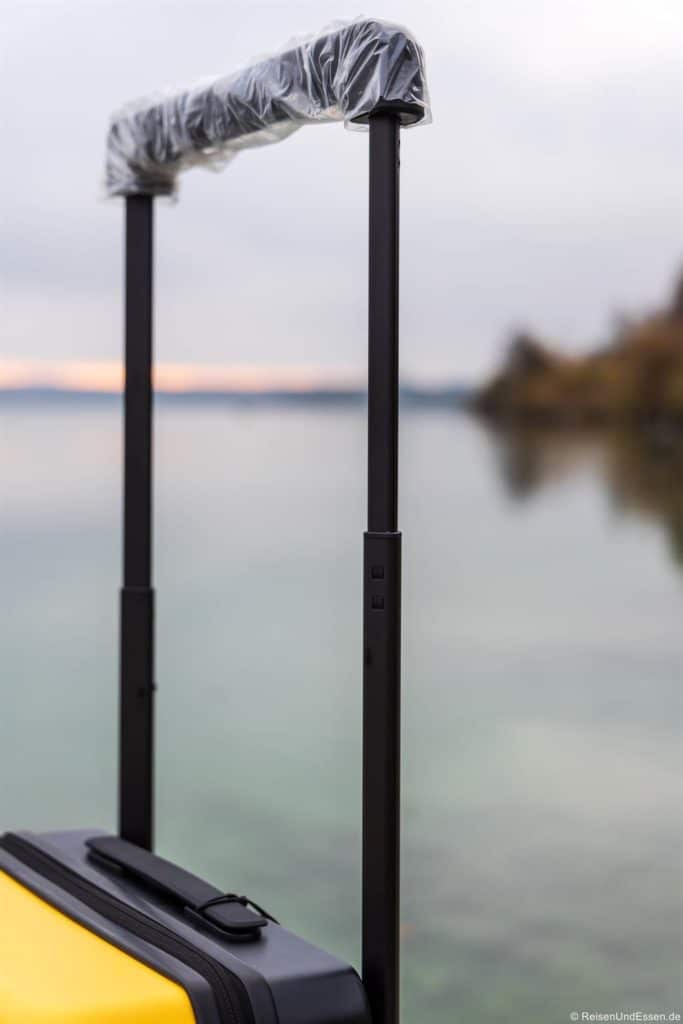Nahaufnahme des verlängerten Griffs eines gelben Koffers, der noch mit Schutzfolie bedeckt ist. Der Hintergrund ist unscharf und zeigt ein ruhiges Gewässer und einen bewölkten Himmel mit einigen undeutlichen Bäumen auf der rechten Seite.