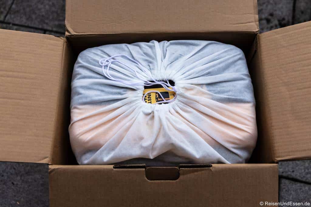 Handgepäck-Koffer für Kurzreisen: Eine Pappschachtel mit geöffneten Klappen gibt den Blick auf einen ordentlich verpackten Level8 Voyageur Koffer frei, der mit einem weißen Kordeltuch bedeckt ist. Ein Teil eines gelben Etiketts ist durch den Stoff in der Mitte sichtbar. Die Schachtel steht auf einer gefliesten Fläche.