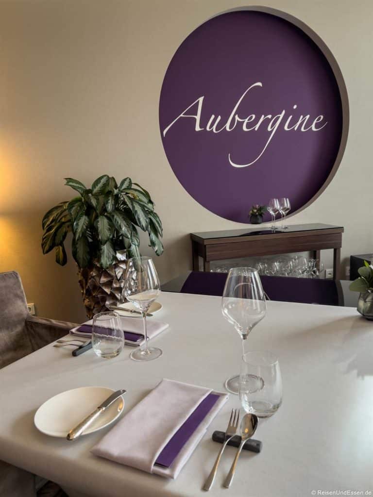 Ein moderner Restauranttisch im Aubergine in Starnberg ist mit weißen und violetten Servietten, Weingläsern, Besteck und einem weißen Teller gedeckt. Hinter dem Tisch stehen eine Pflanze und eine Vitrine mit Gläsern unter einem großen lilafarbenen Wandkreis, auf dem in Schreibschrift das Wort Aubergine steht.