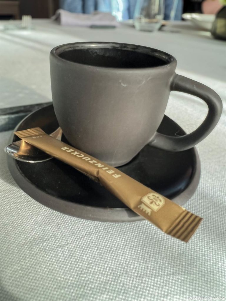 Eine mattschwarze Kaffeetasse steht auf einer passenden Untertasse, auf der ein Löffel und ein goldenes Zuckerpäckchen mit der Aufschrift Feinzucker ruhen. Die Tasse ist leer, und die Szene ist hell beleuchtet und wirft weiche Schatten auf ein helles Tischtuch.