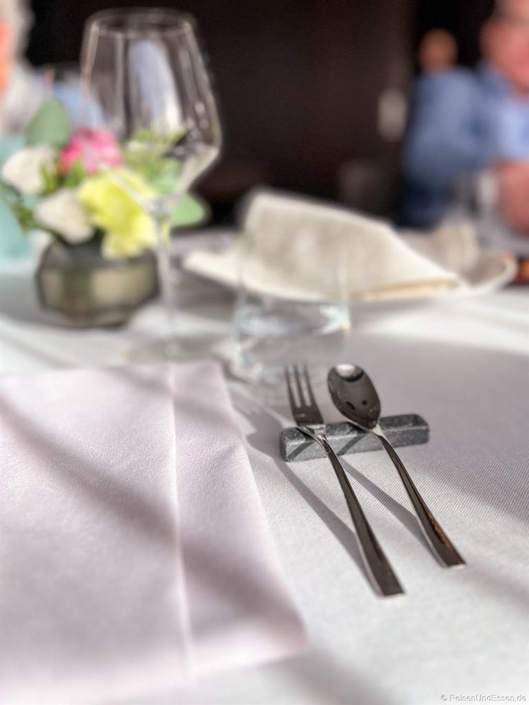 Eine Nahaufnahme eines gedeckten Tisches im Aubergine Starnberg mit einer gefalteten weißen Serviette, einer Gabel und einem Löffel, die auf einer dunklen Stäbchenablage ruhen, einem Weinglas, einem Wasserglas und einem kleinen Blumenarrangement im Weichzeichner im Hintergrund. Das Sonnenlicht wirft sanfte Schatten auf das weiße Tischtuch.