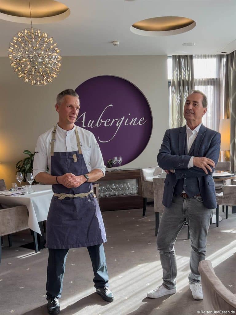 Zwei Männer stehen in einem eleganten Restaurant. Max Moder links trägt eine Chefkoch-Uniform mit blauer Schürze, die Hände ineinander verschränkt. Armin Langer rechts, gekleidet in einen grauen Blazer und ein weißes Hemd, steht selbstbewusst mit verschränkten Armen da. Hinter ihnen steht ein lila Schild mit der Aufschrift "Aubergine".