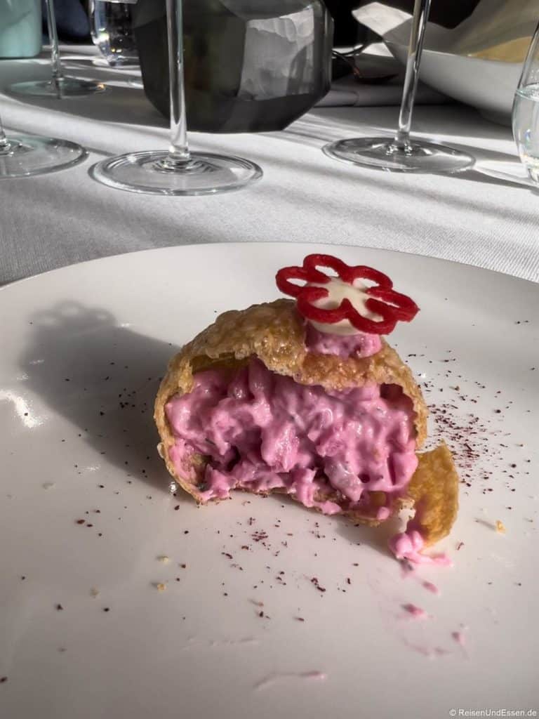 Eine mit cremigem rosa Salat gefüllte Teighülle wird auf einem weißen Teller aufgeschnitten. Die Füllung ist stückig und lebendig. Das Gebäck ist mit einer dünn geschnittenen, dekorativen rot-weißen Blume verziert. Im Hintergrund sind Weingläser und Geschirr auf einem weißen Tischtuch zu sehen.