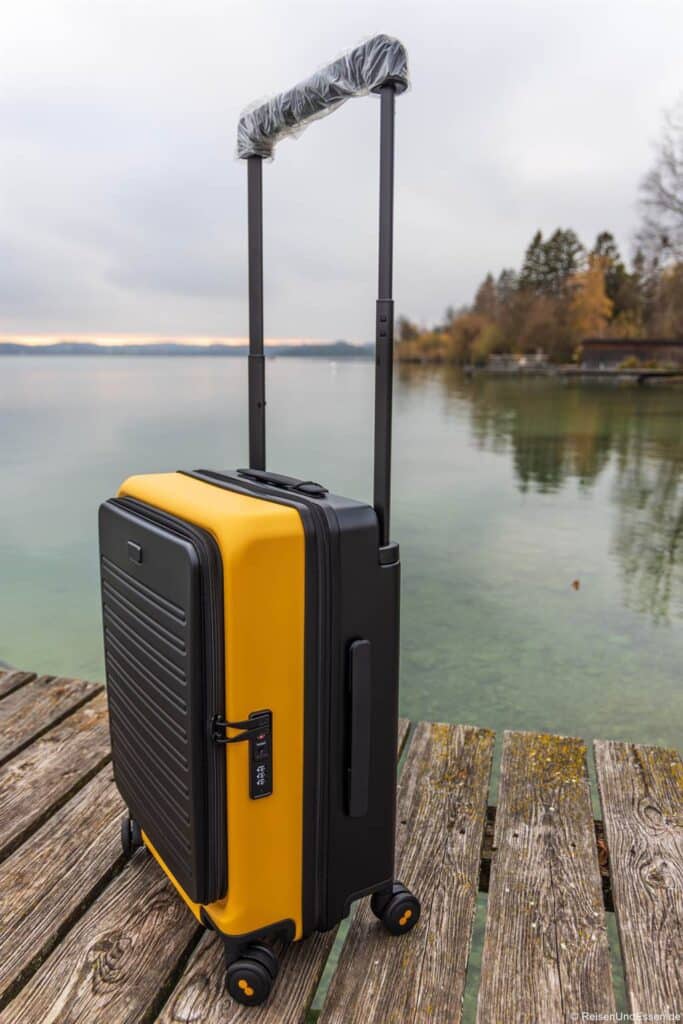 Ein gelb-schwarzer Hartschalenkoffer mit vier Rädern und einem kunststoffummantelten Griff steht auf einem verwitterten Holzsteg an einem ruhigen See, mit Bäumen und bewölktem Himmel im Hintergrund.