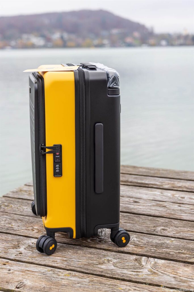 Ein gelb-schwarzer Hartschalenkoffer mit vier Rädern steht auf einem hölzernen Steg an einem ruhigen See, mit einem nebelverhangenen, baumbestandenen Hügel im Hintergrund unter einem bewölkten Himmel. An der Seite des Koffers befindet sich ein eingebautes Zahlenschloss.