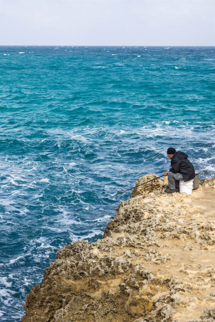 Eine Person in dunkler Jacke und Hut sitzt auf einer felsigen Klippe und angelt mit einer Angel über den leuchtend türkisfarbenen, kabbeligen Meereswellen. Der Himmel ist blass und die Felsen sind zerklüftet und kontrastieren mit dem leuchtenden Meer unter ihnen.