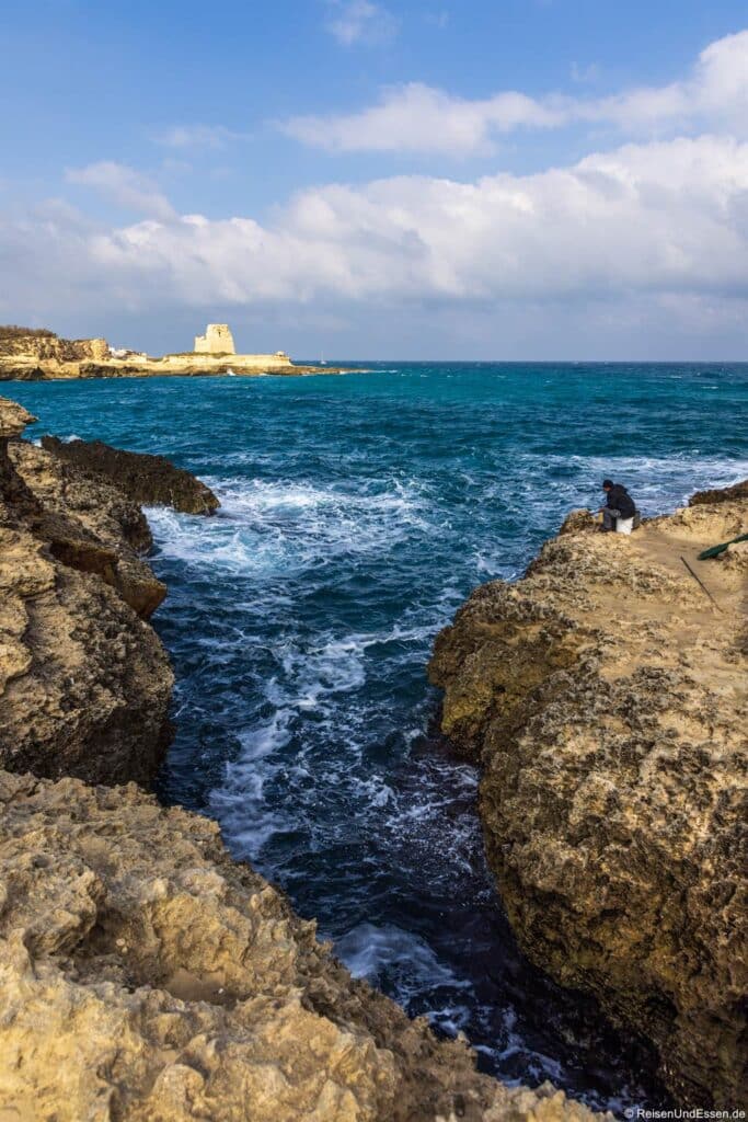 Grotta della Poesia: Eine Person sitzt auf zerklüfteten, felsigen Klippen an einer schmalen Bucht mit kabbeligem, blaugrünem Wasser. In der Ferne steht ein alter Steinturm auf einer kleinen Landzunge unter einem teilweise bewölkten Himmel, und an der Küste sind weitere Felsen zu sehen.