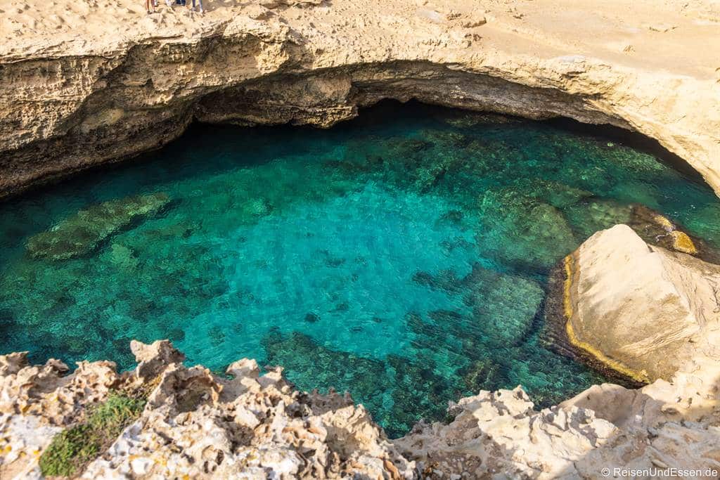Die Grotta della Poesia mit klarem türkisfarbenem Wasser, umgeben von zerklüfteten beigen Felsen. Das Sonnenlicht beleuchtet das leuchtend blaugrüne Wasser und lässt die Felsen und Schatten unter der Wasseroberfläche erkennen. Die felsigen Ränder rahmen den Pool ein und schaffen eine abgelegene, ruhige Szene.